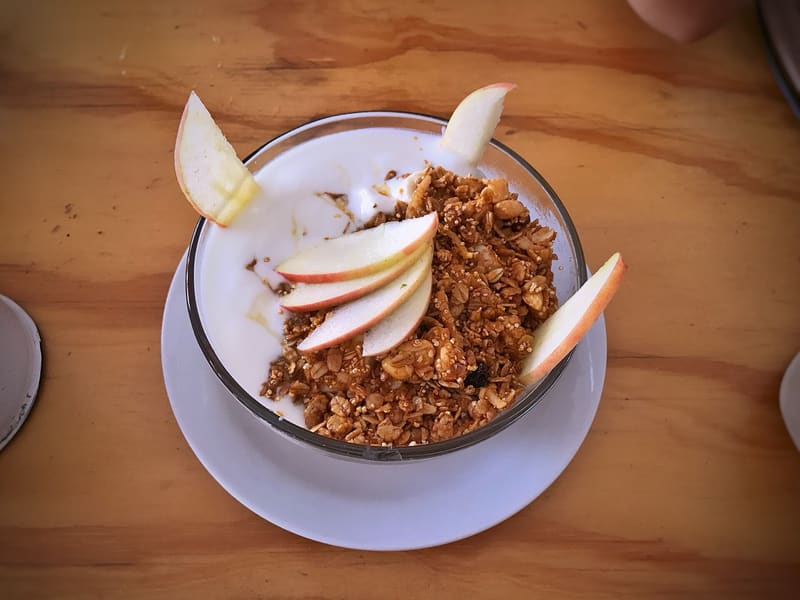 Overhead view of a glass bowl of yogurt topped with granola and fanned apple slices, on a white saucer on a wooden table