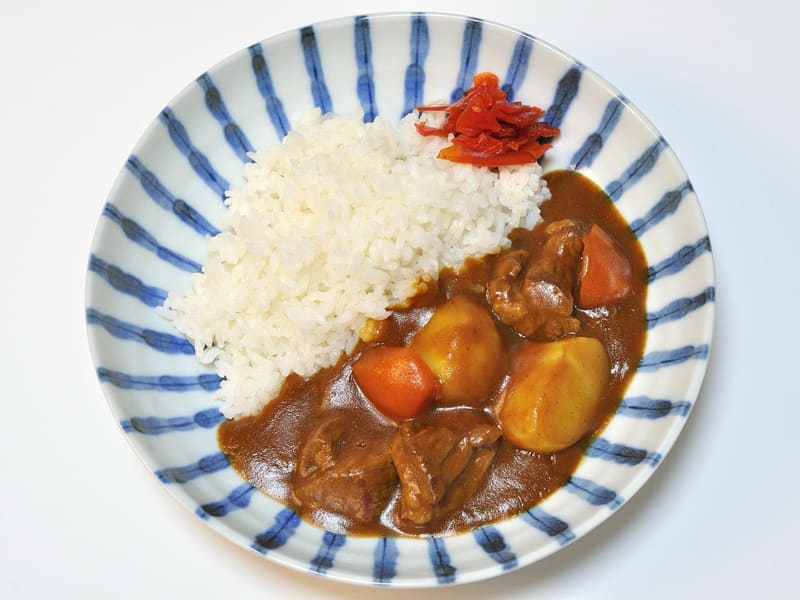 Overhead view of Japanese beef curry with white rice, potato and carrot in a blue and white striped bowl, with a small pile of red pickle on the rim