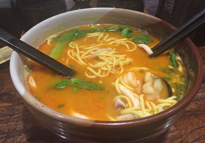 Bowl of satay noodles in an orange peanut broth with yellow noodles, green bok choy, shrimp and squid, black chopsticks resting in the bowl
